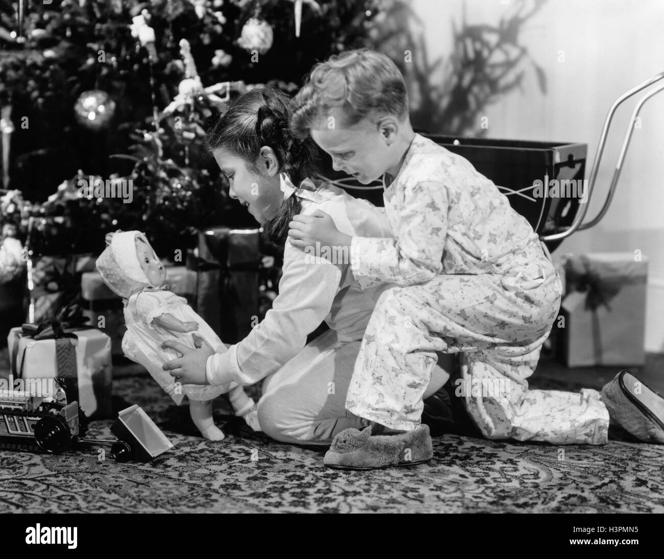 1940s BOY GIRL BROTHER SISTER LOOKING AT PRESENTS UNDER CHRISTMAS TREE GIRL HOLDING BABY DOLL Stock Photo
