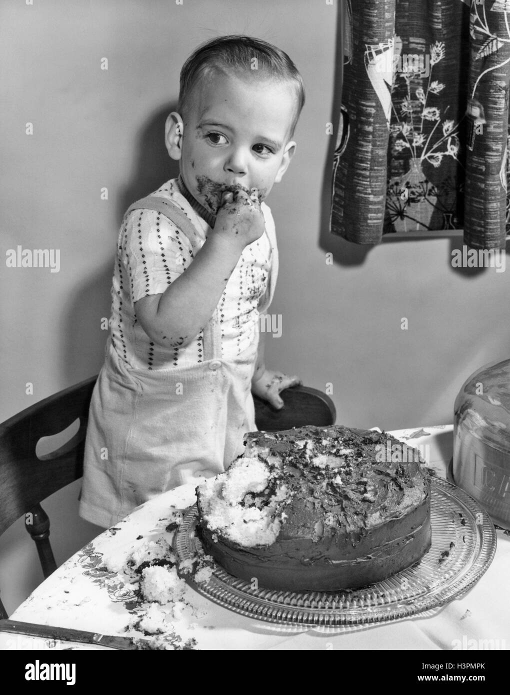 1950s LITTLE BOY TODDLER STANDING ON CHAIR BY CHOCOLATE CAKE WITH FINGERS IN MOUTH AND ICING ALL OVER HIS FACE Stock Photo