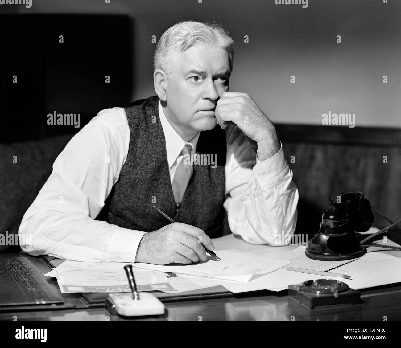 1920s 1930s WORRIED BUSINESSMAN WEARING VEST AND SHIRT SLEEVES SITTING AT DESK THINKING ABOUT SIGNING A DOCUMENT Stock Photo