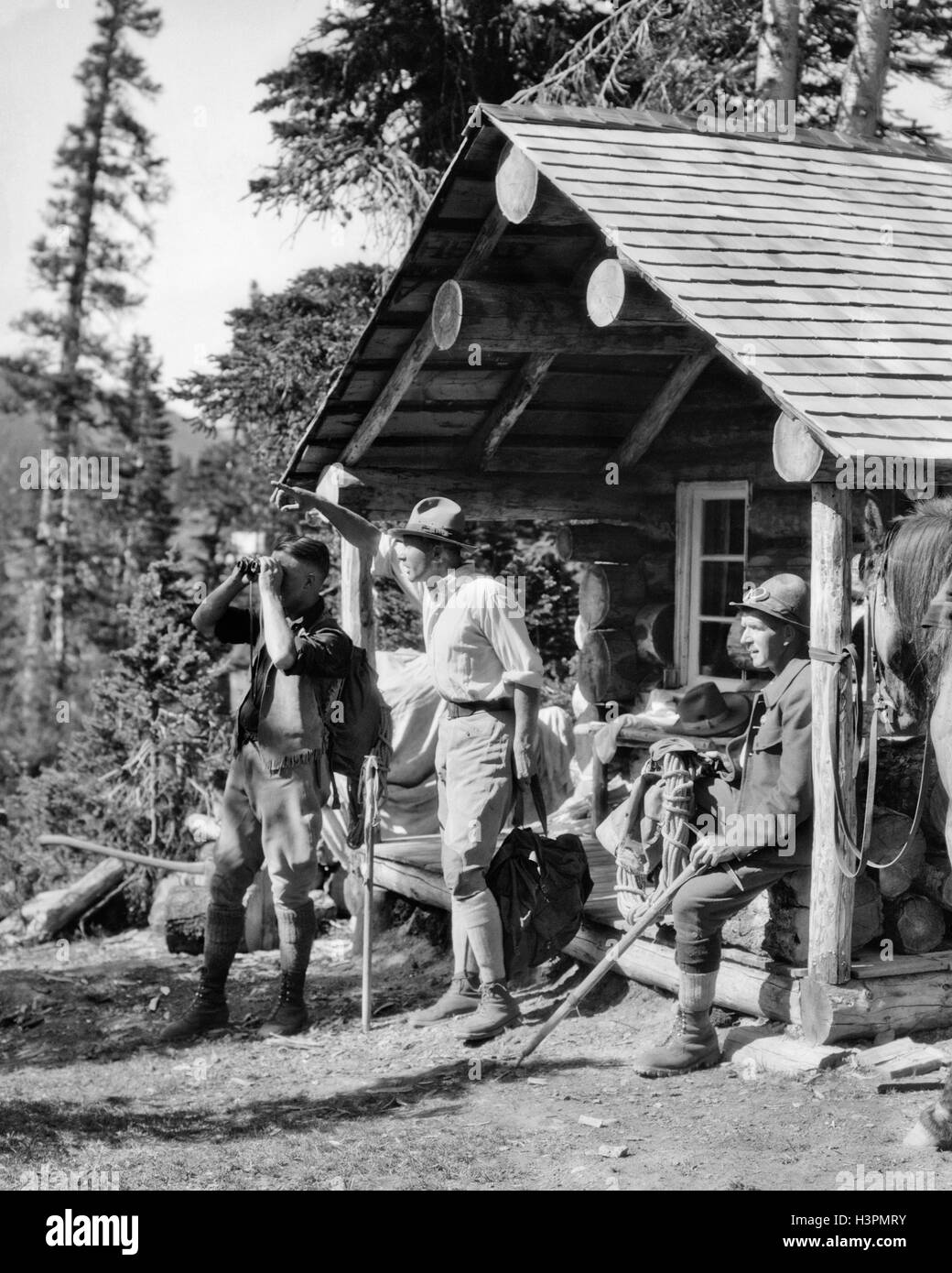 1920s THREE MEN BY PORCH OF LOG CABIN CAMP WITH HORSE ONE MAN USING BINOCULARS OTHER MAN POINTING ASSINIBOINE CANADA Stock Photo