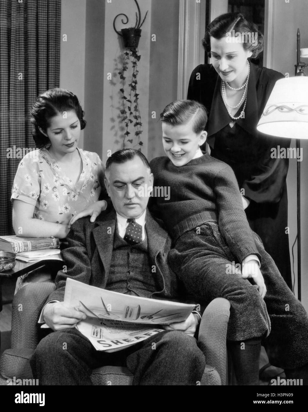 1940s FAMILY OF FOUR WOMAN MOTHER TEENAGE DAUGHTER GIRL AND SON BOY SITTING AROUND FATHER MAN READING NEWSPAPER Stock Photo
