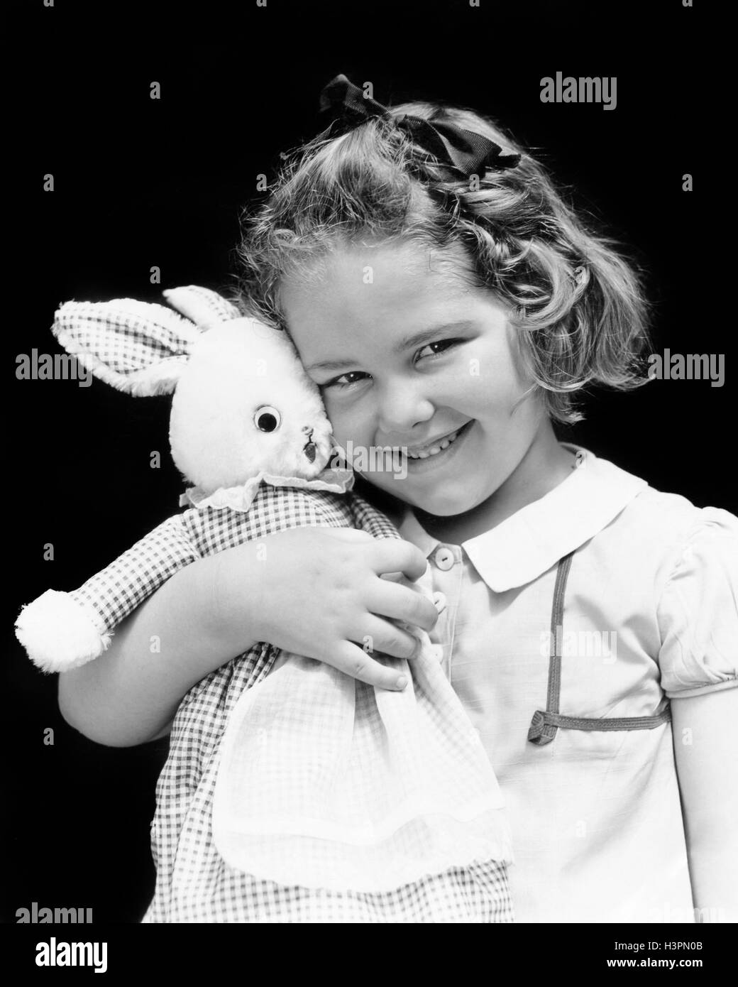 1930s 1940s SMILING YOUNG GIRL LOOKING AT CAMERA HOLDING HUGGING STUFFED RABBIT TOY ANIMAL Stock Photo