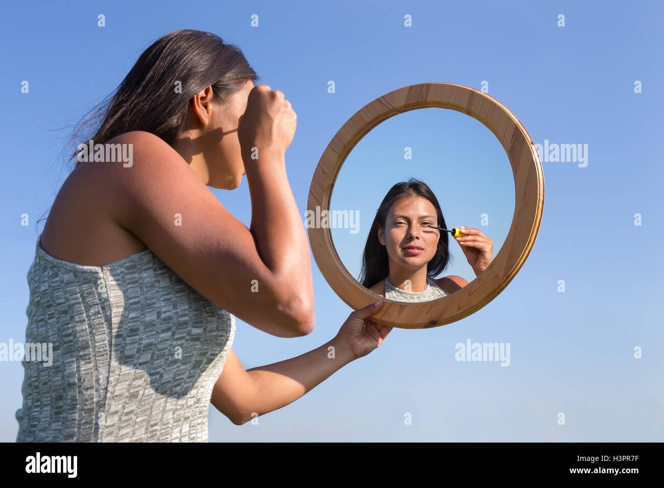 Woman applying cosmetics mascara makeup in mirror outside with blue sky