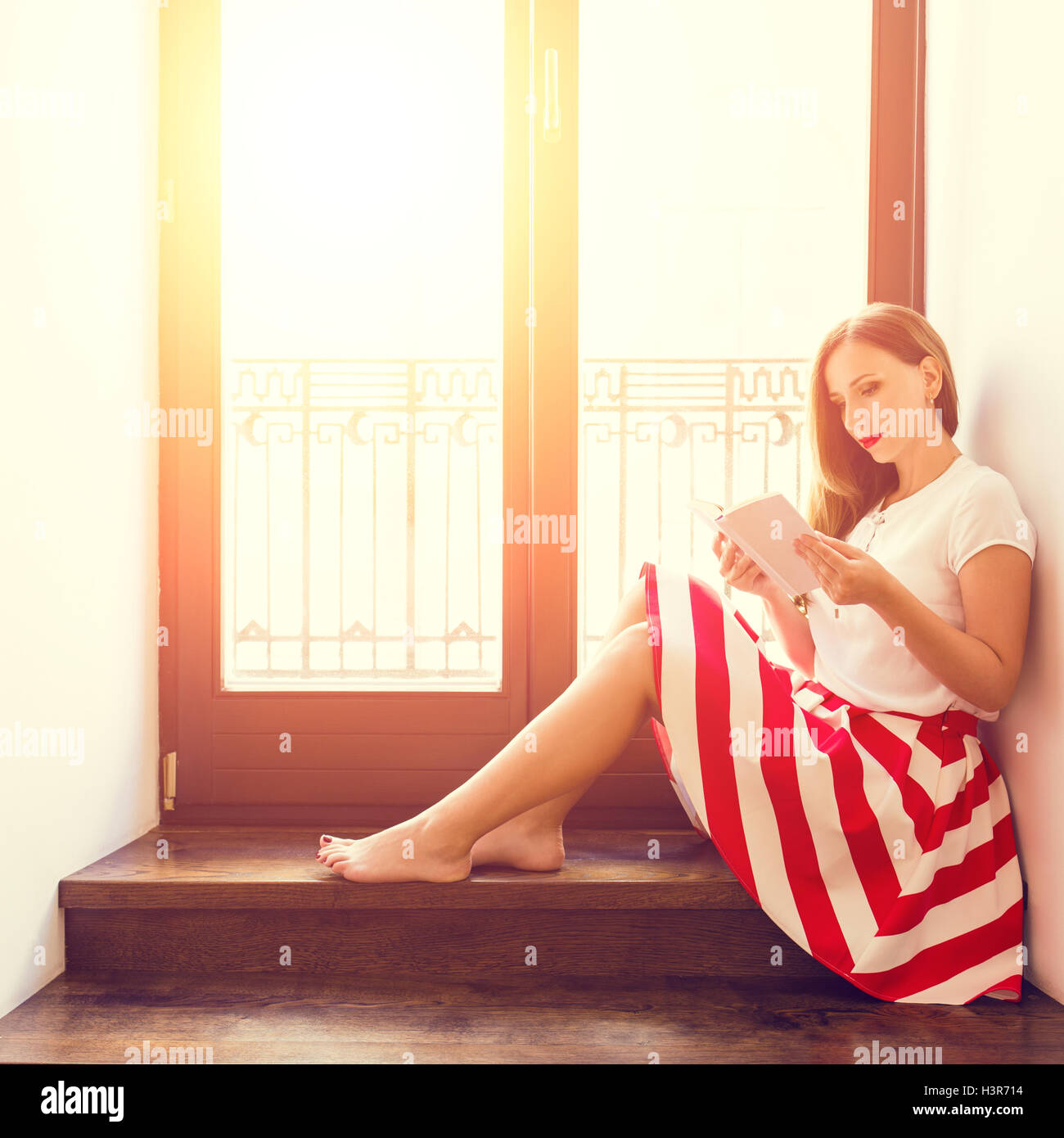 Young stylish lady reading book near balcony glass door. Beautiful girl with book on wooden stairs in hotel Stock Photo