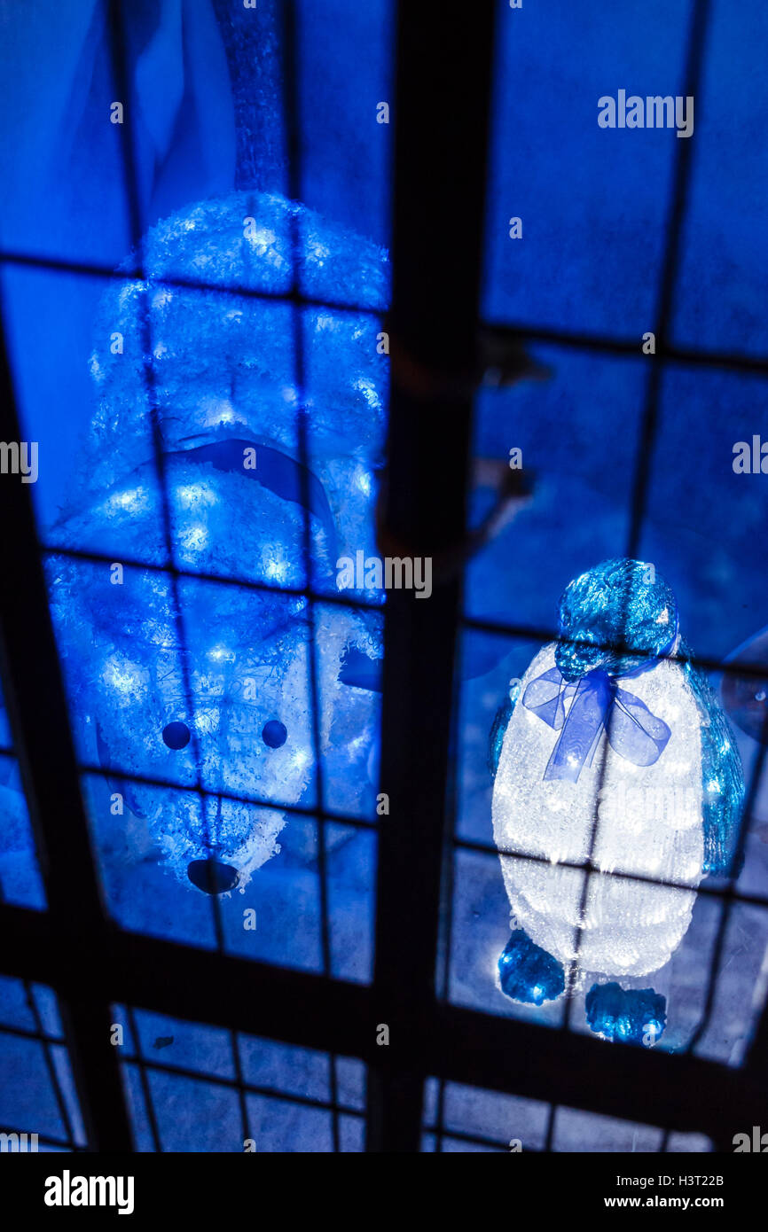 Blue illuminated polar bear and penguin Christmas ornaments behind wire mesh, as if imprisoned Stock Photo