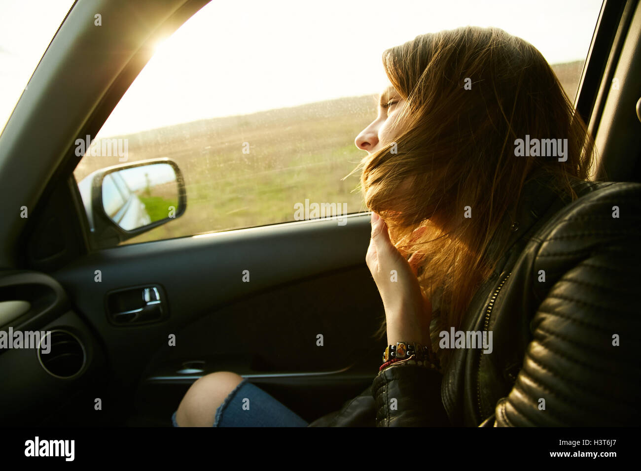 A girl in a leather jacket is looking in the window of a car at a field while travelling Stock Photo