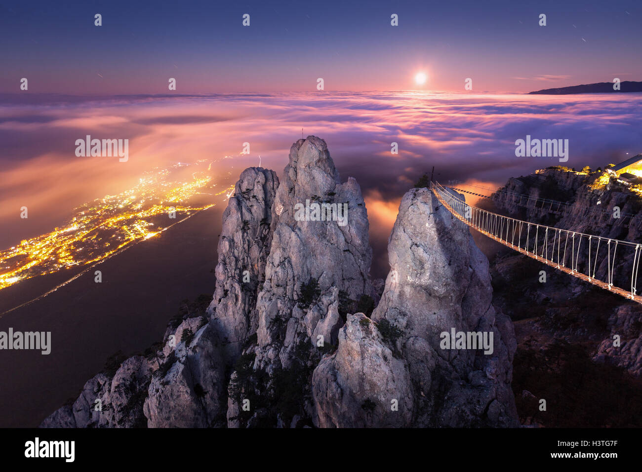 Mountain landscape with rising full moon and fog in the city at night. Beautiful night landscape with mountain peak, bridge, low Stock Photo