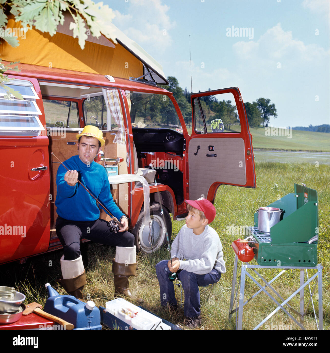 1970s MAN AND BOY CAMPING SON SITTING BY PORTABLE STOVE FATHER SITTING BY CAMPER WITH FISHING RODS AND GEAR Stock Photo