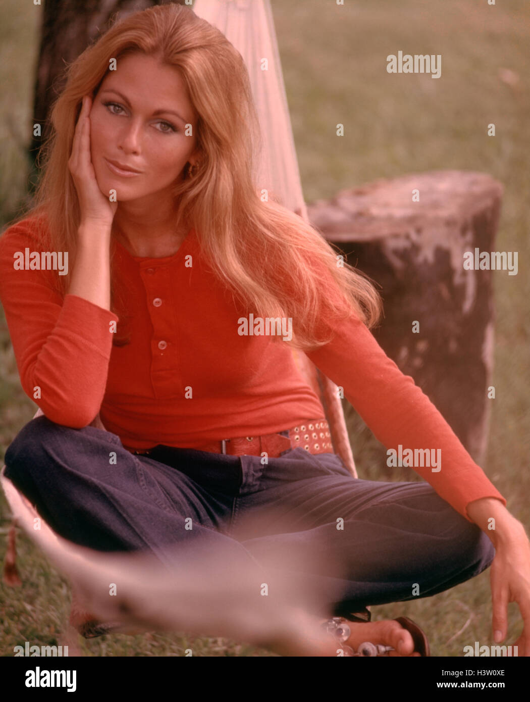 1970s SMILING SEXY WOMAN LONG BLOND HAIR SITTING OUTDOORS RED TEE SHIRT BELT HIP HUGGER BLUE SLACKS Stock Photo