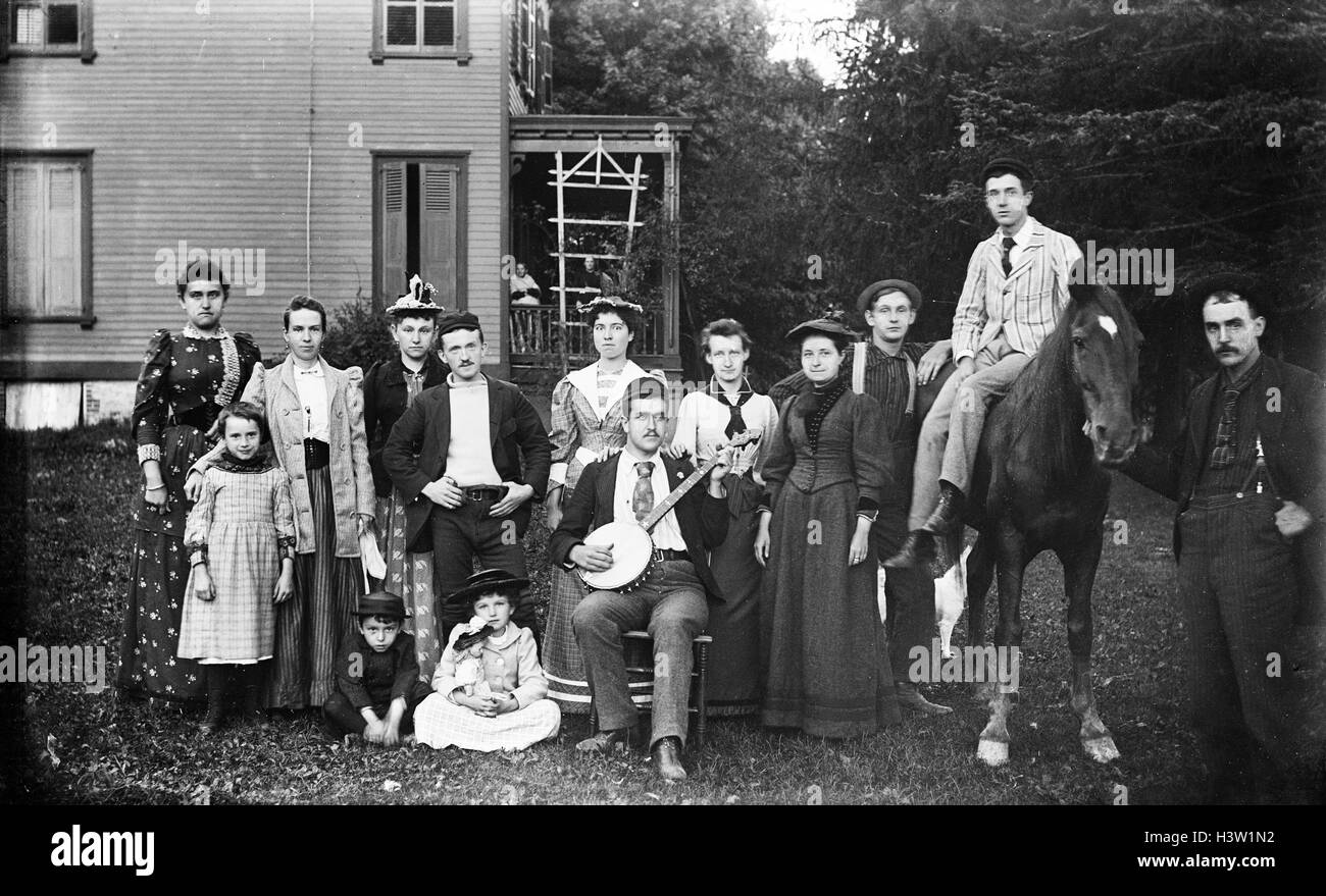 1890s GROUP PORTRAIT FAMILY LOOKING AT CAMERA POSING ON LAWN IN FRONT OF HOUSE MAN IN CENTER HOLDING BANJO MAN SITTING ON HORSE Stock Photo