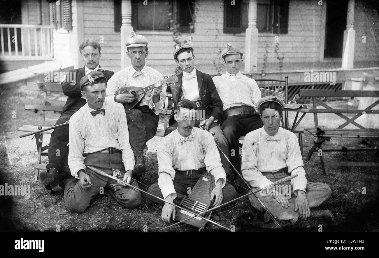 1900s GROUP PORTRAIT MEN POSING WITH MUSICAL INSTRUMENTS MANDOLIN AUTOHARP AND FISHING RODS SITTING ON LAWN IN FRONT OF HOUSE Stock Photo