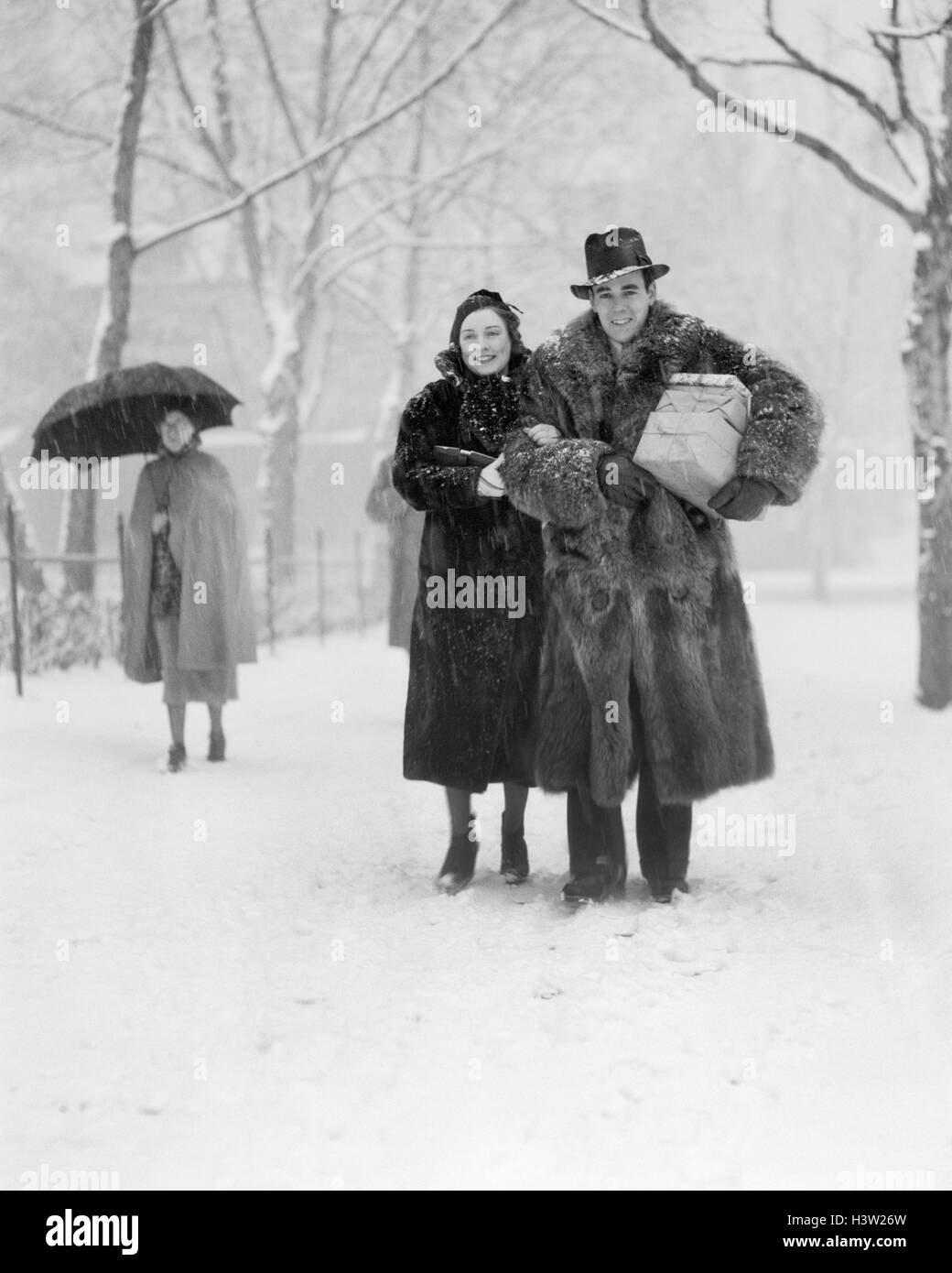 1930s SMILING CHRISTMAS SHOPPING COUPLE WEARING FUR COATS WALKING THROUGH SNOWY CITY PARK CARRYING PACKAGES LOOKING AT CAMERA Stock Photo