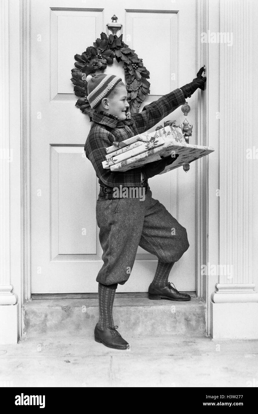 1930s 1940s BOY HOLDING WRAPPED PACKAGES RINGING BELL ON DOOR WITH CHRISTMAS WREATH Stock Photo