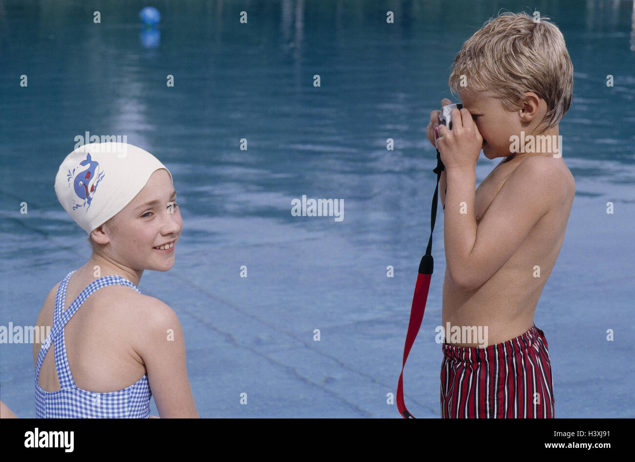 Pool, cymbal margin, girl, sit, boy, take of a photo, detail, model released, outside, swimming-pool, swimming pool, swimming pool, summer, leisure time, holidays, outdoor swimming pool, siblings, swimming cap, camera, photo camera, camera, side view Stock Photo