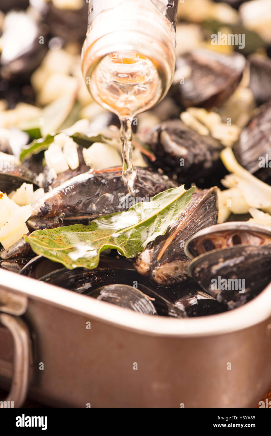 Pouring white wine on clams. Seafood dish preparation. Rustic gourmet dinner with mussels, onion and bay leaf. Stock Photo