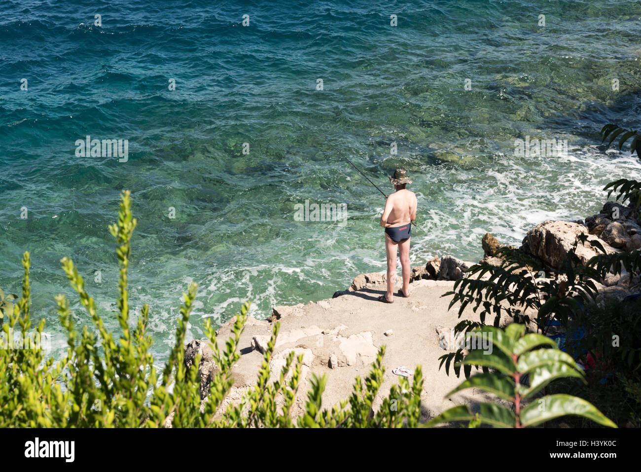 A man wearing swimming trunks fishing on a rocky seashore at Trpanj Croatia on a sunny summer day Stock Photo