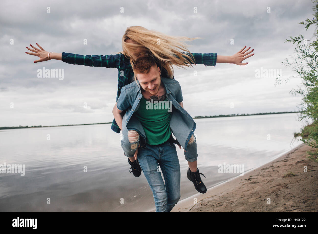 Happy man piggybacking girlfriend on lakeshore against sky Stock Photo