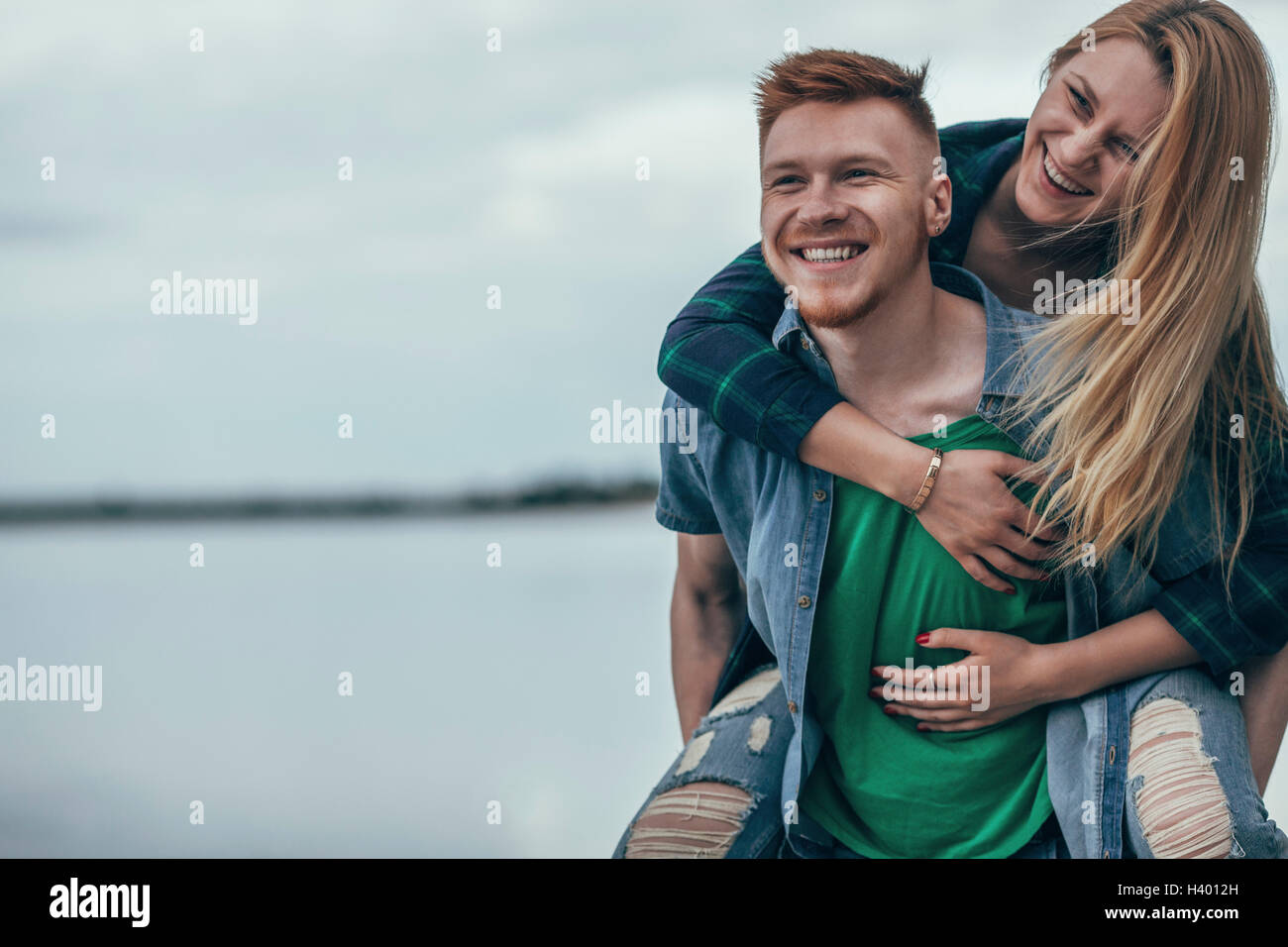 Happy man piggybacking cheerful girlfriend on lakeshore against sky Stock Photo