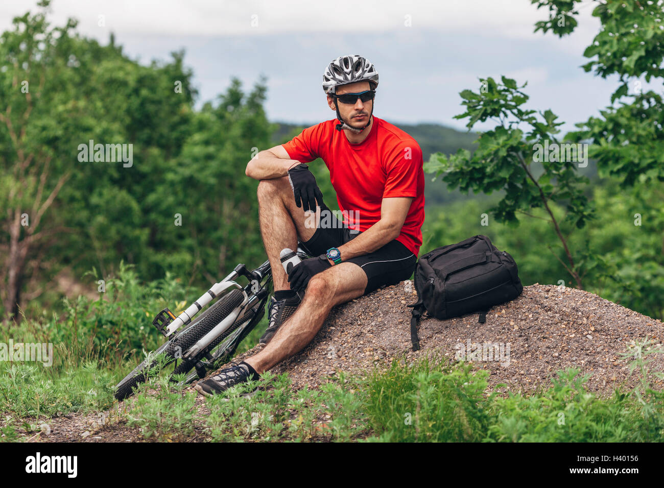 Tired mountain biker sitting on ground against trees Stock Photo