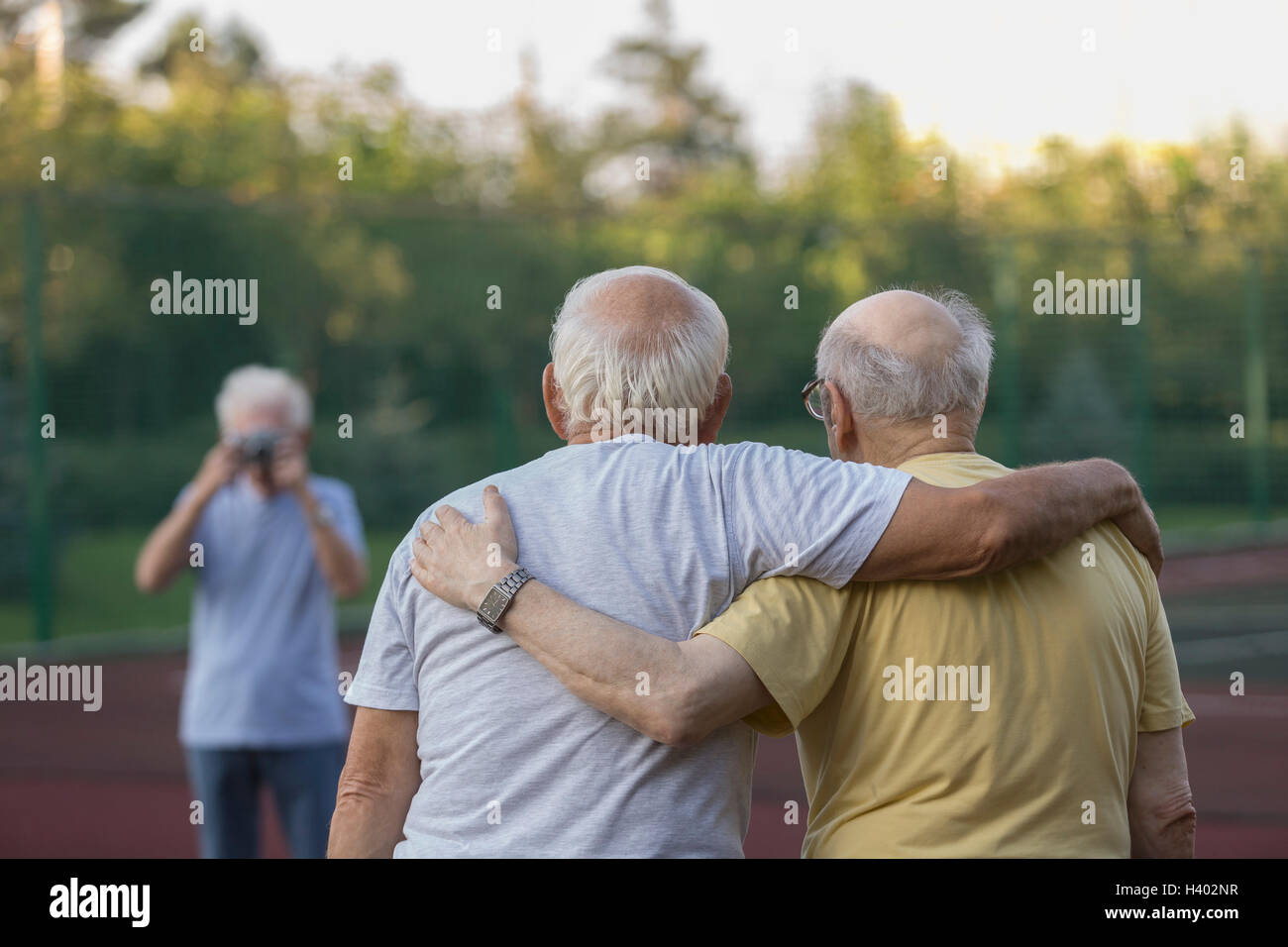 Rear view of friends standing with arm around while man photographing in background Stock Photo