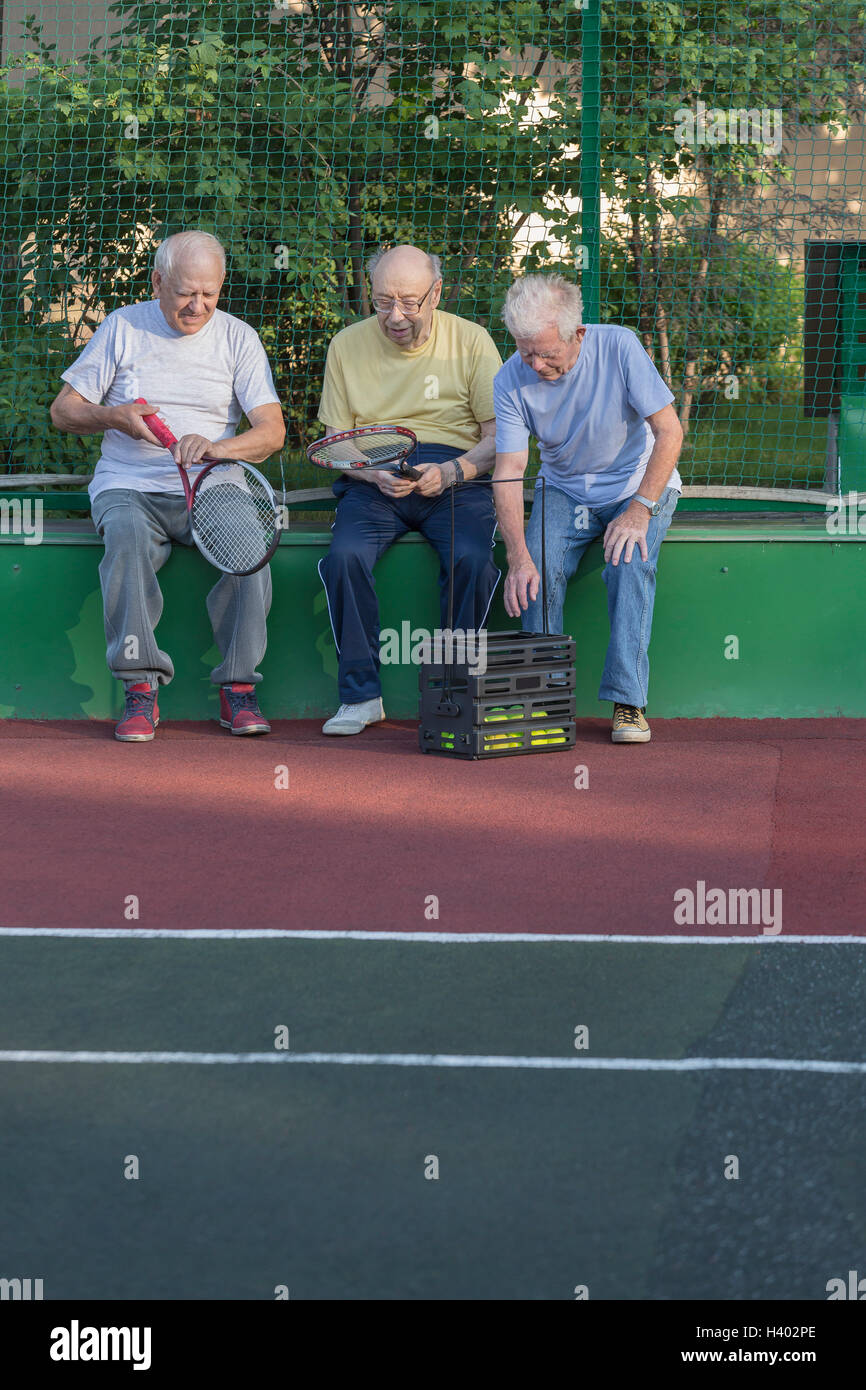 Senior friends with tennis rackets sitting against fence at playing field Stock Photo
