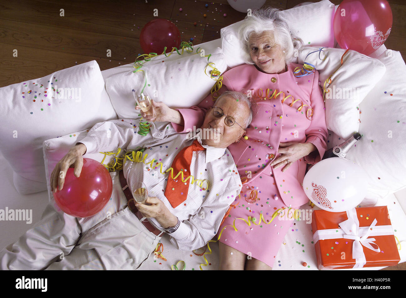 Sofa, senior pair, tipsily, confetti,  Balloons, gift, streamers,  from above Pair, seniors, pensioners, party, celebration, birthday celebration, ceremony, celebrates, together, relationship, partnership, affection, love, falls in love, happily, smiles, exhaustion, pause, rests, lies, champagne glasses, birthday, tenderness Stock Photo