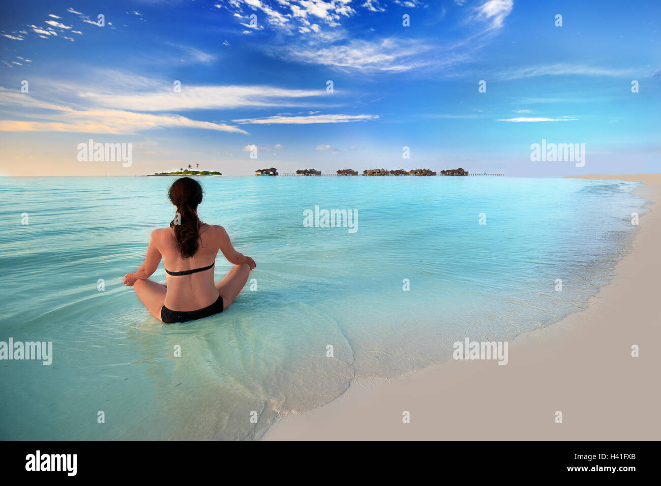 Young woman exercising yoga in turquoise lagoon with overwater bungalows on tropical island Stock Photo
