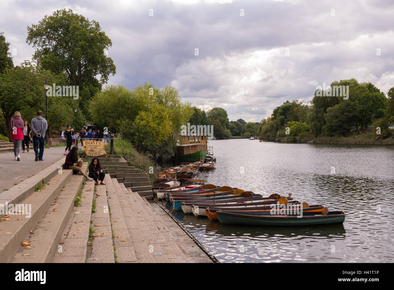 People walking or sitting down enjoying the warm day next to the river Thames on Richmond riverfront with multiple rowing boats Stock Photo