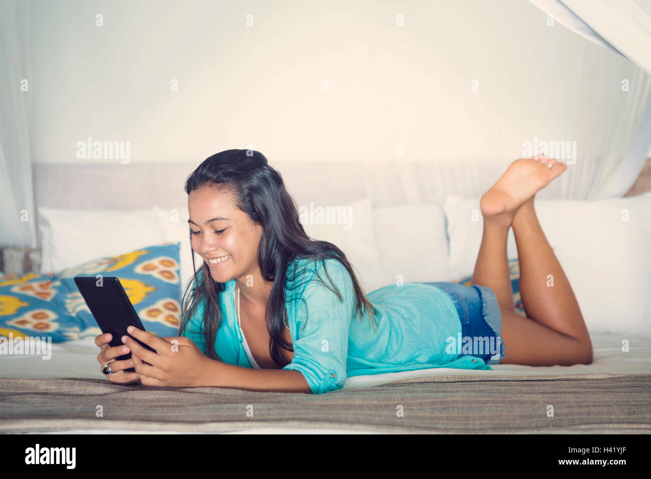 Mixed Race girl laying on bed reading digital tablet Stock Photo