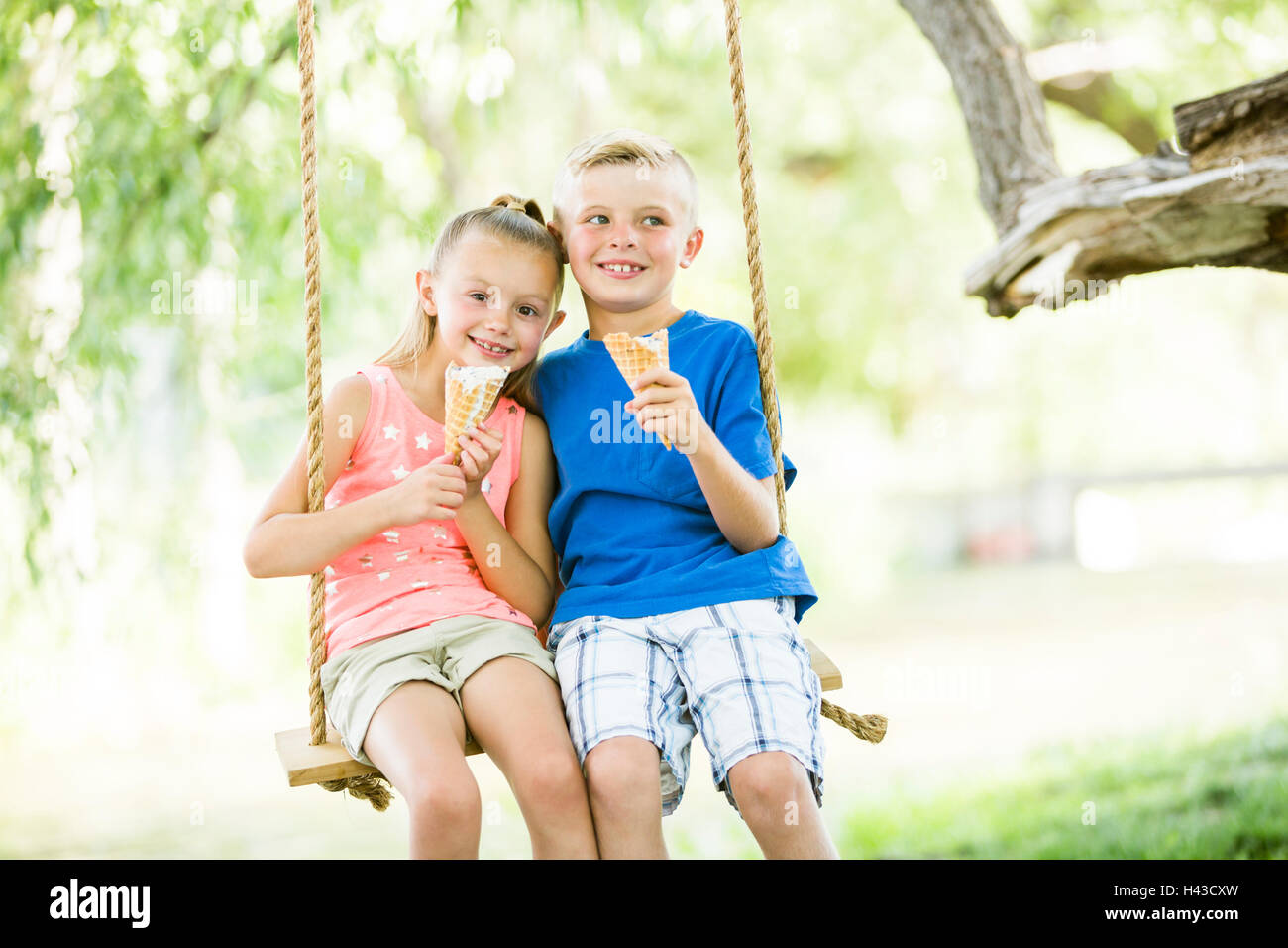 Caucasian brother and sister eating ice cream cones on rope swings Stock Photo