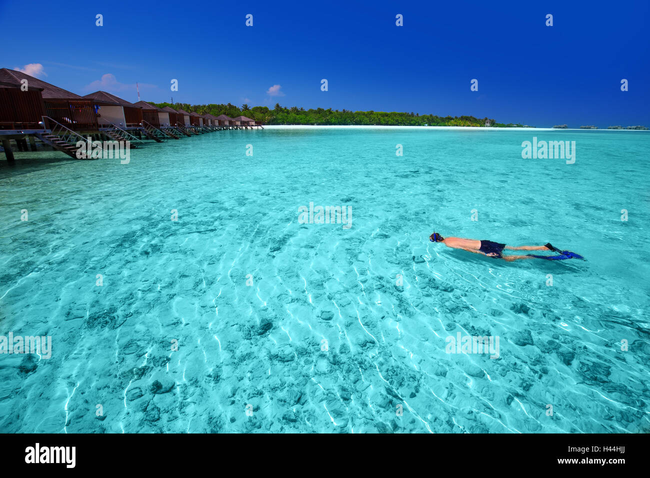 Young man snorkeling in tropical island with sandy beach, palm trees, overwater bungalows and turquoise clear water Stock Photo