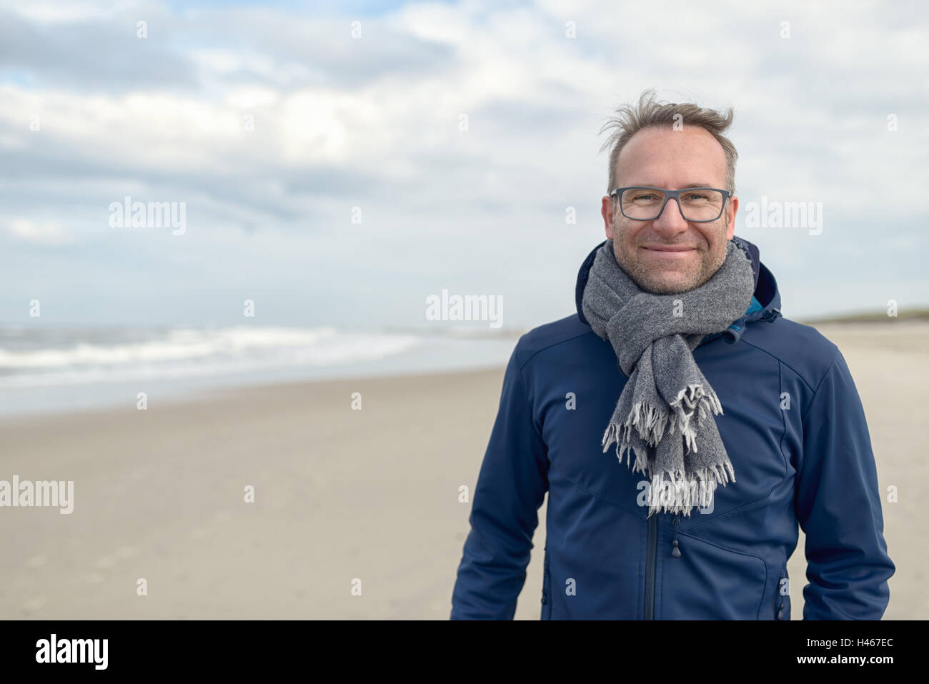 Smiling middle-aged man wearing glasses and a knitted woollen scarf standing on a deserted autumn beach on a cloudy day with cop Stock Photo