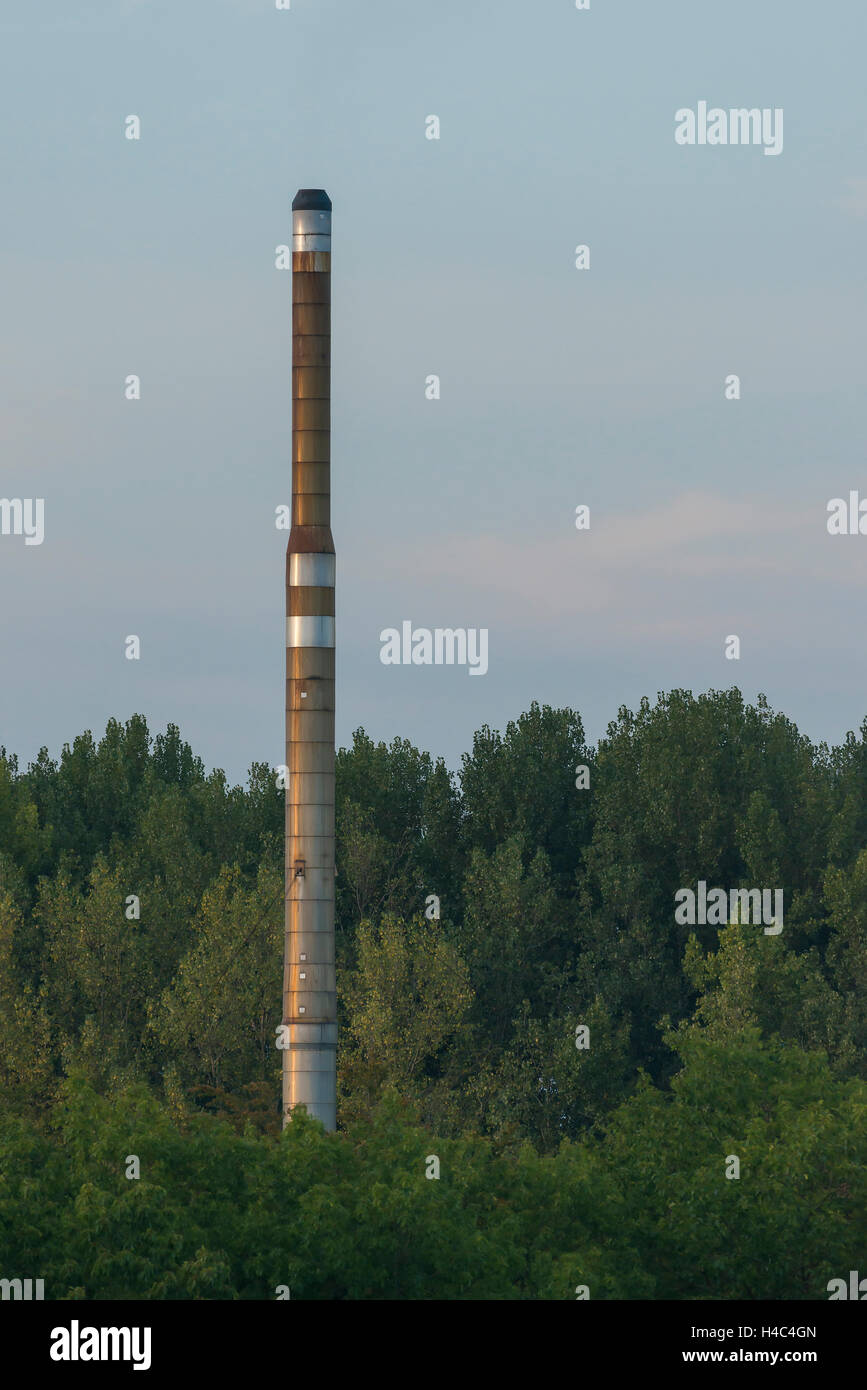 Factory chimney of a factory located in a forested area visible from a nearby higher point in the evening light Stock Photo