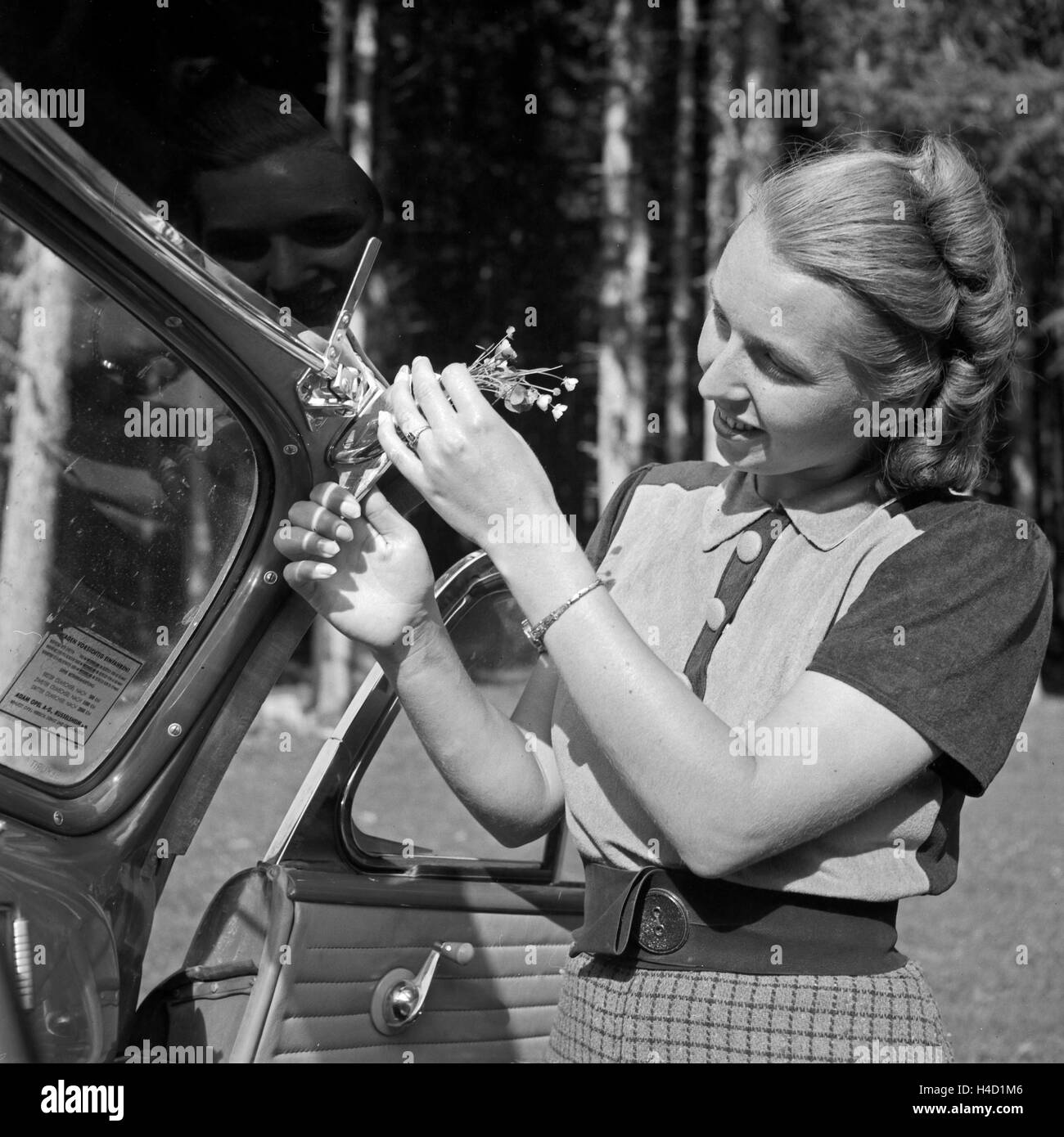 Eine junge Frau befestigt einen kleinen Blumengruß an der Windschutscheibe, Österreich 1930er Jahr. A young woman sticking a few flowers to the screen of a car, Germany 1930s. Stock Photo