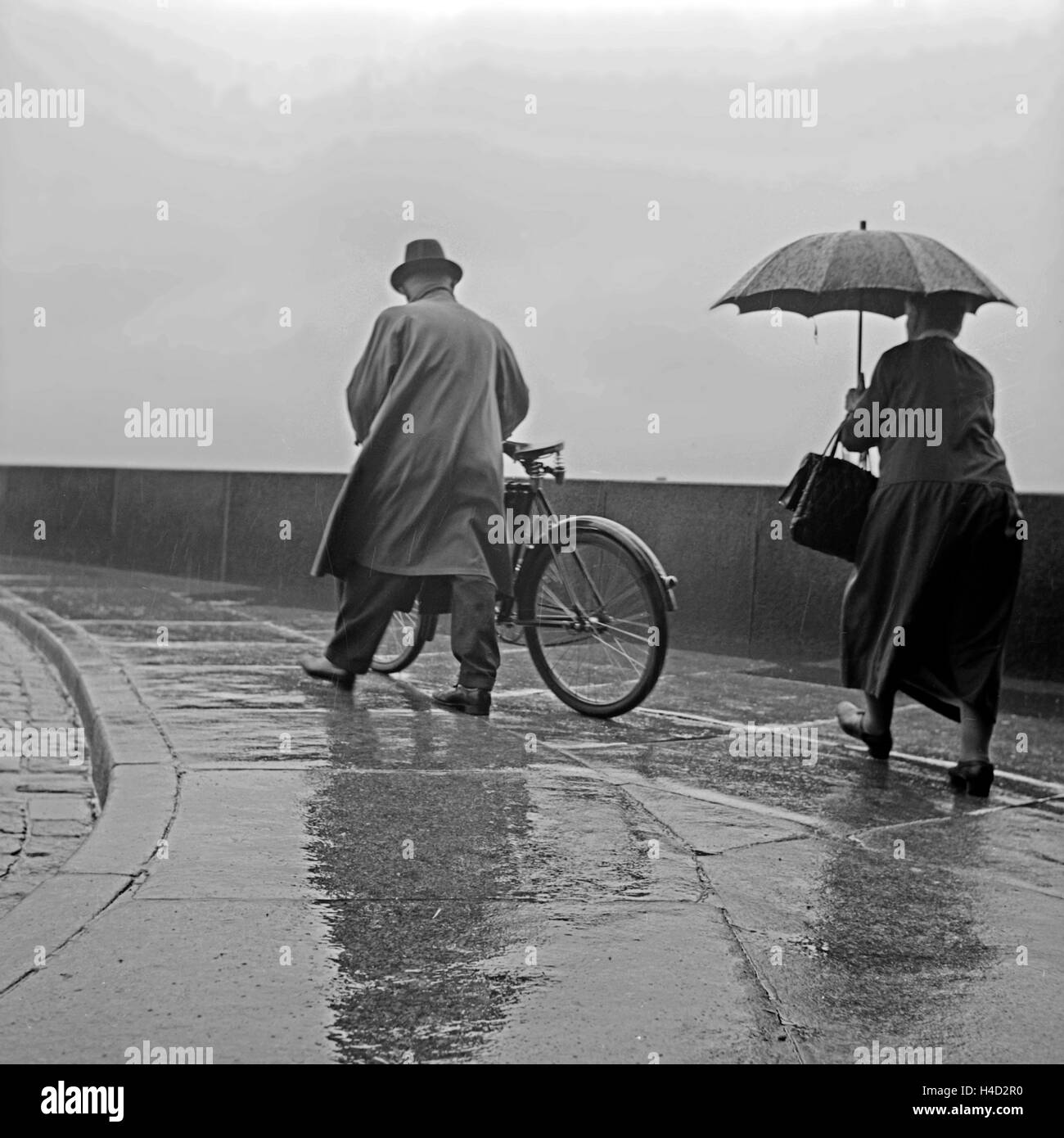 Ein Mann schiebt sein Fahrrad durch den Regen, eine Frau folgt ihm mit ihrem Regenschirm, Deutschland 1930er Jahre. A man pushing his bicycle, a woman following him with her umbrella in the rain, Germany 1930s. Stock Photo