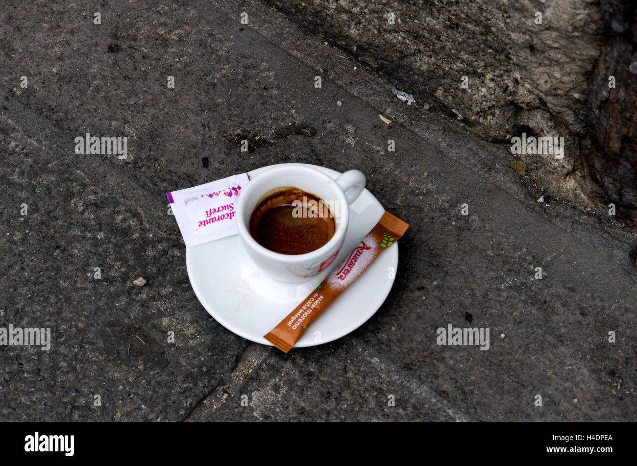 Partially finished cup of espresso is left on pavement by edge of stone wall. Two unopened packets of sugar on the saucer. Stock Photo