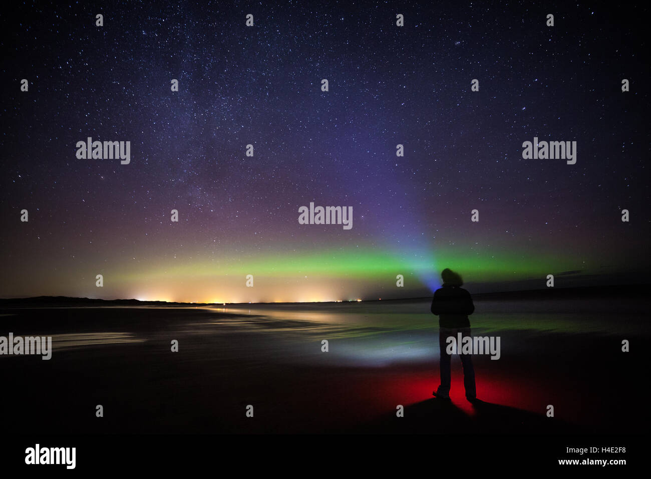 A lone woman shining a torch up at the night sky with the Milky Way and Aurora Borealis, the Northern Lights, from the beach Stock Photo