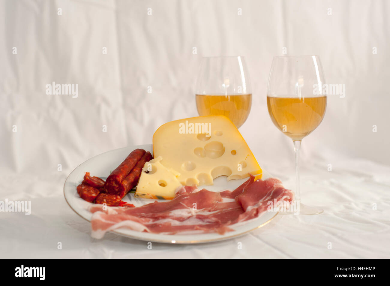 Plate of meats and cheese appetizer with two stemmed wine glasses with white wine on white cloth Stock Photo