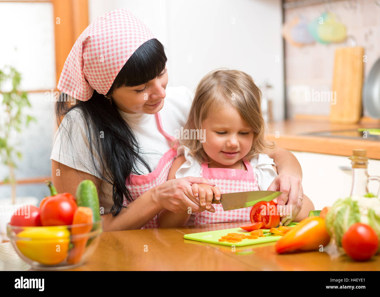 Mother teaching child making salad in kitchen. Mom and kid chopping vegetable on cutting board with knife. Cooking concept of happy family preparing healthy food for dinner. Stock Photo