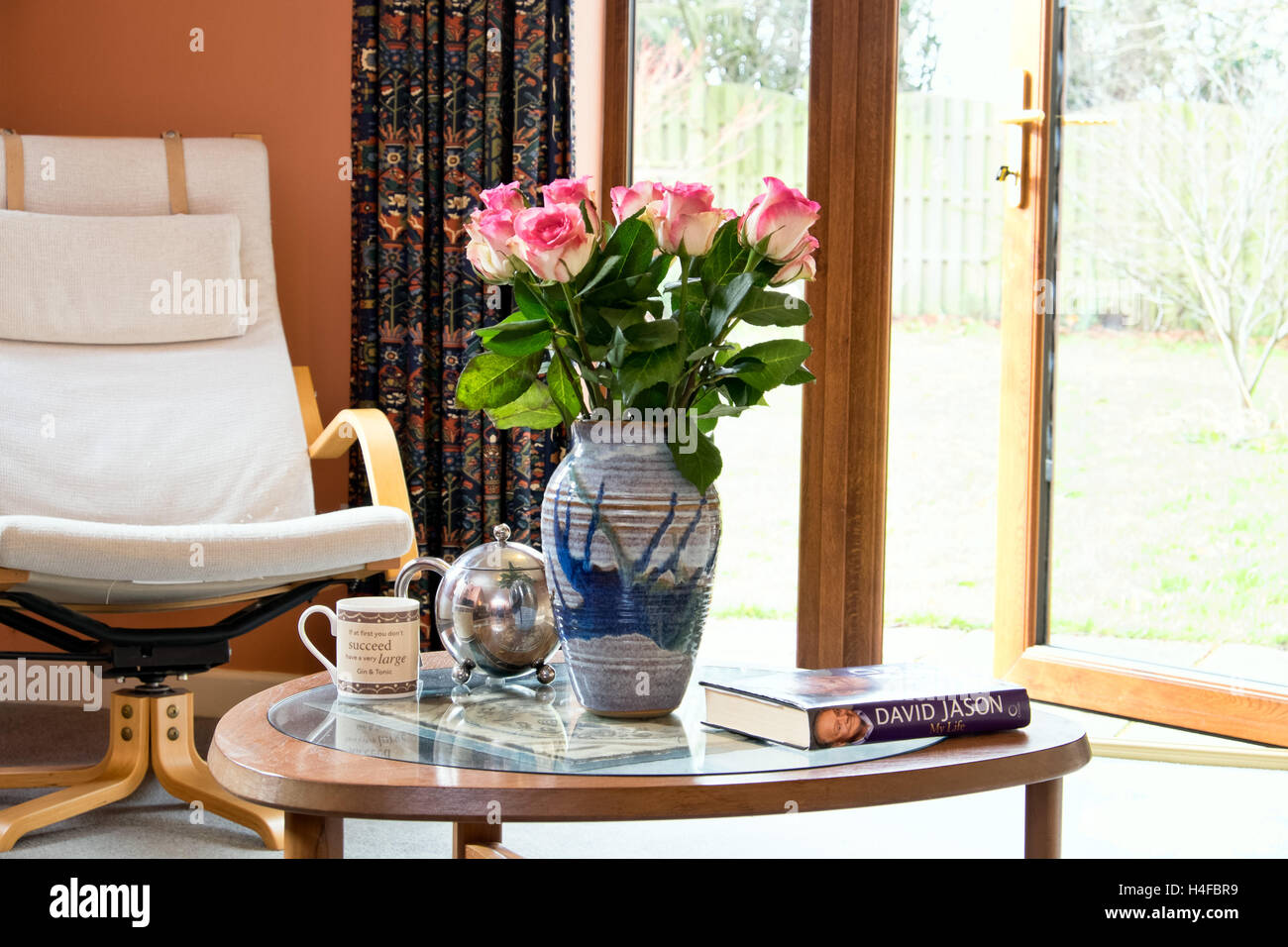 A lifestyle image of a coffee table with roses, teapot, mug & book in an average UK home with a garden outlook. Stock Photo