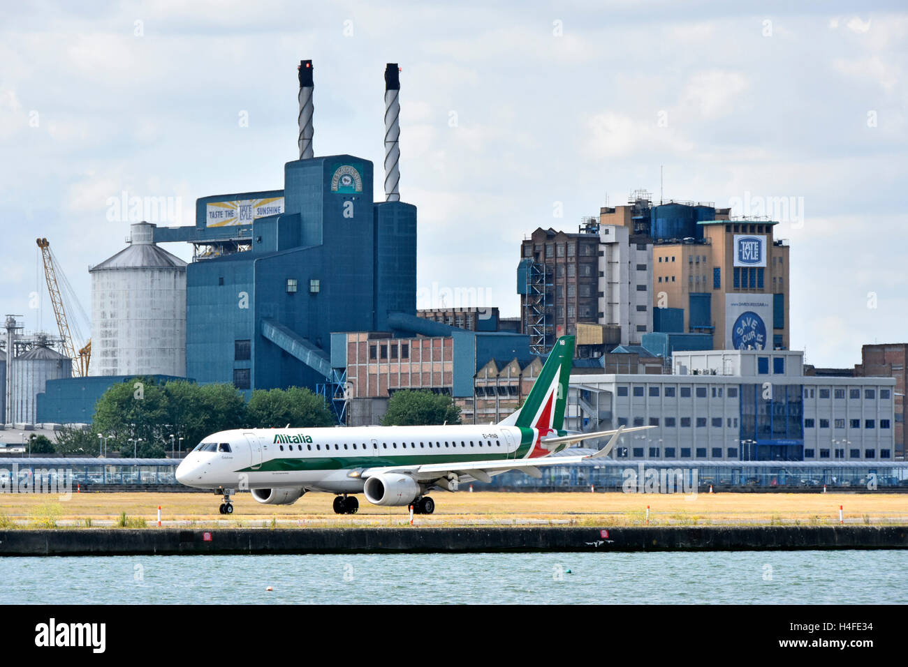 Airplane Alitalia Embraer taxiing before take off London City Airport Tate & Lyle Silvertown sugar refinery factory buildings beyond Newham England UK Stock Photo