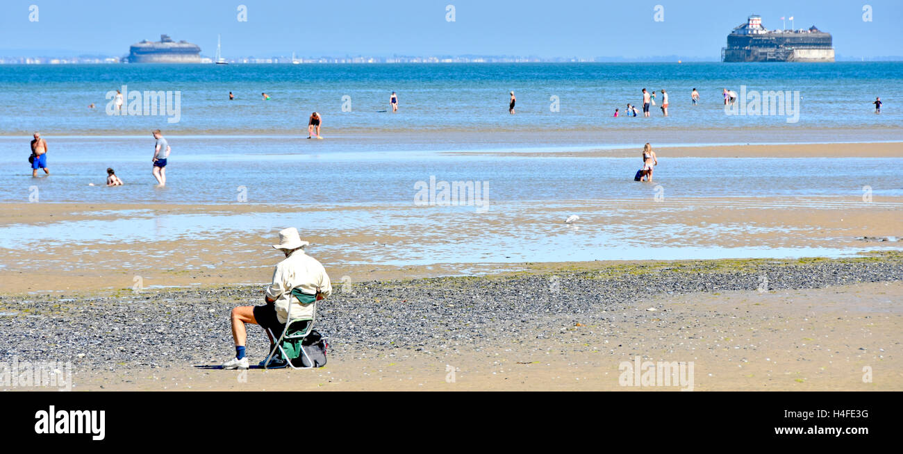 Back view older man sitting alone on Isle of Wight UK holiday beach at low tide sketching views of the Solent including two forts on hot summer day Stock Photo