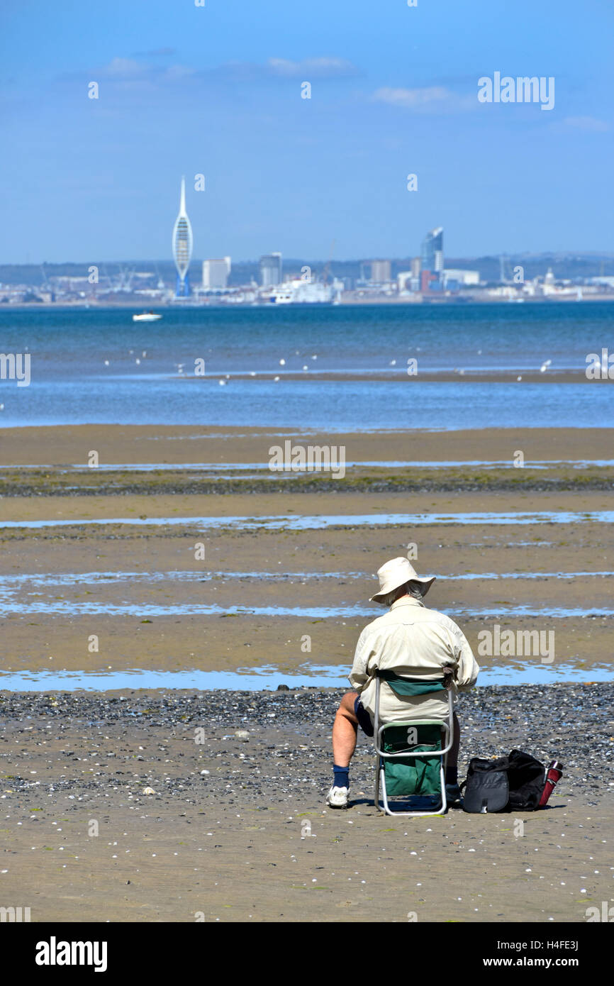 Older man sitting alone on Isle of Wight UK seaside beach at low tide sketching views of the Solent towards distant Portsmouth on a hot summer day Stock Photo