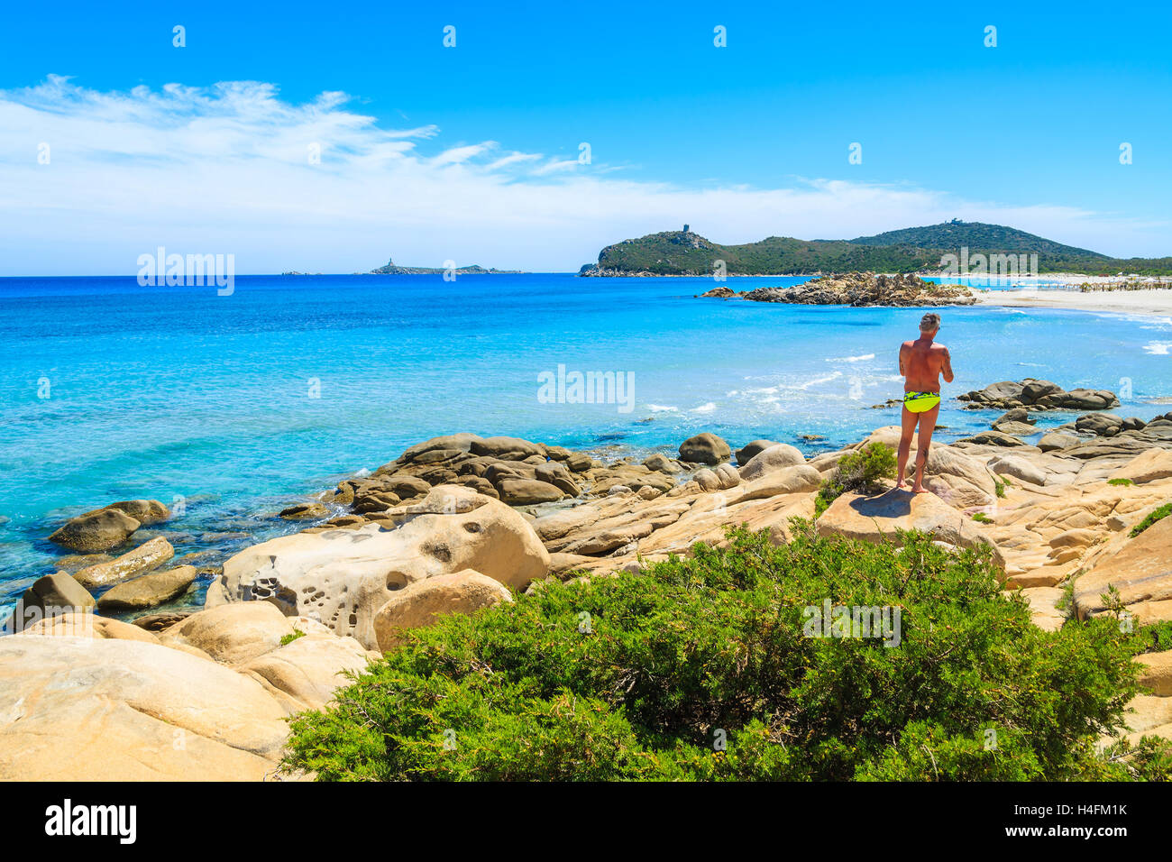 PORTO GIUNCO BAY, SARDINIA ISLAND - MAY 27, 2014: man tourist standing on a rock looking at turquoise sea water on coast of Sardinia island, Villasimius peninsula, Italy. Stock Photo