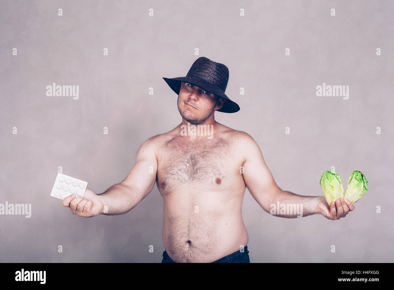 Naked corpulent man in hat giving pharmaceutical products and lettuce. Stock Photo