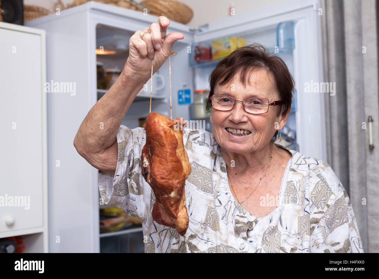 Happy senior woman holding smoked meat while standing in front of the open fridge in the kitchen. Stock Photo