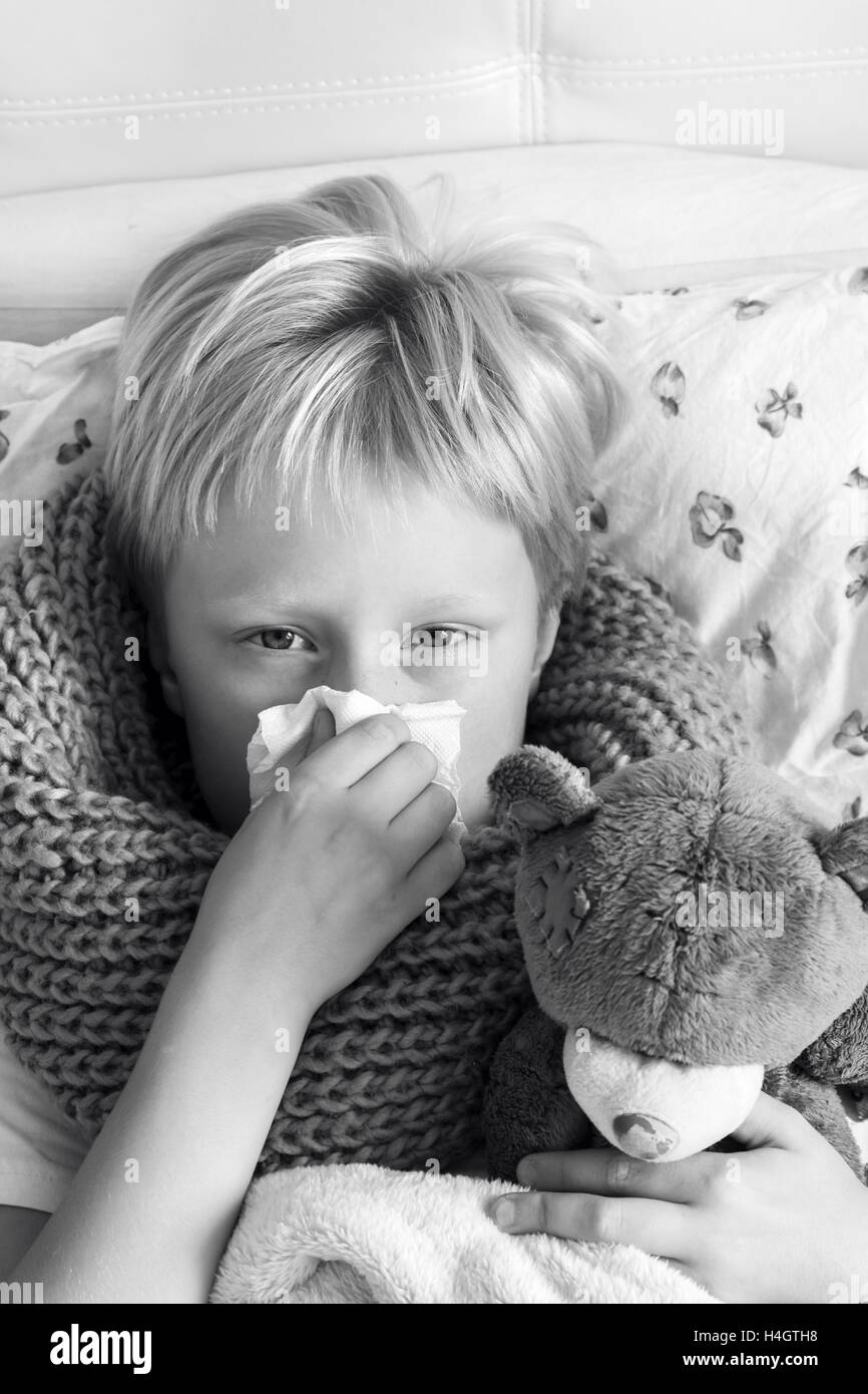 Sick child (blond boy, kid) lying in bed with teddy bear and blowing his nose Stock Photo