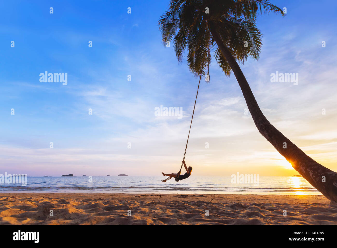 Young happy woman relaxing on a swing attached to a palm tree on a paradise beach at sunset while on vacation in tropics Stock Photo