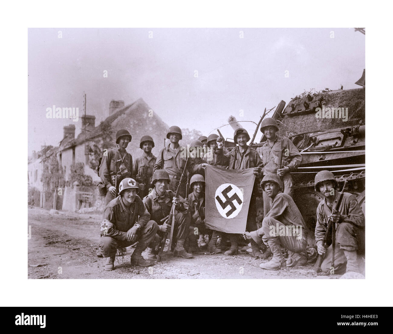 WW2 1945 Northern France American troops celebrate holding swastika flag with capture of German Nazi tank and occupied French village Normandy France Stock Photo