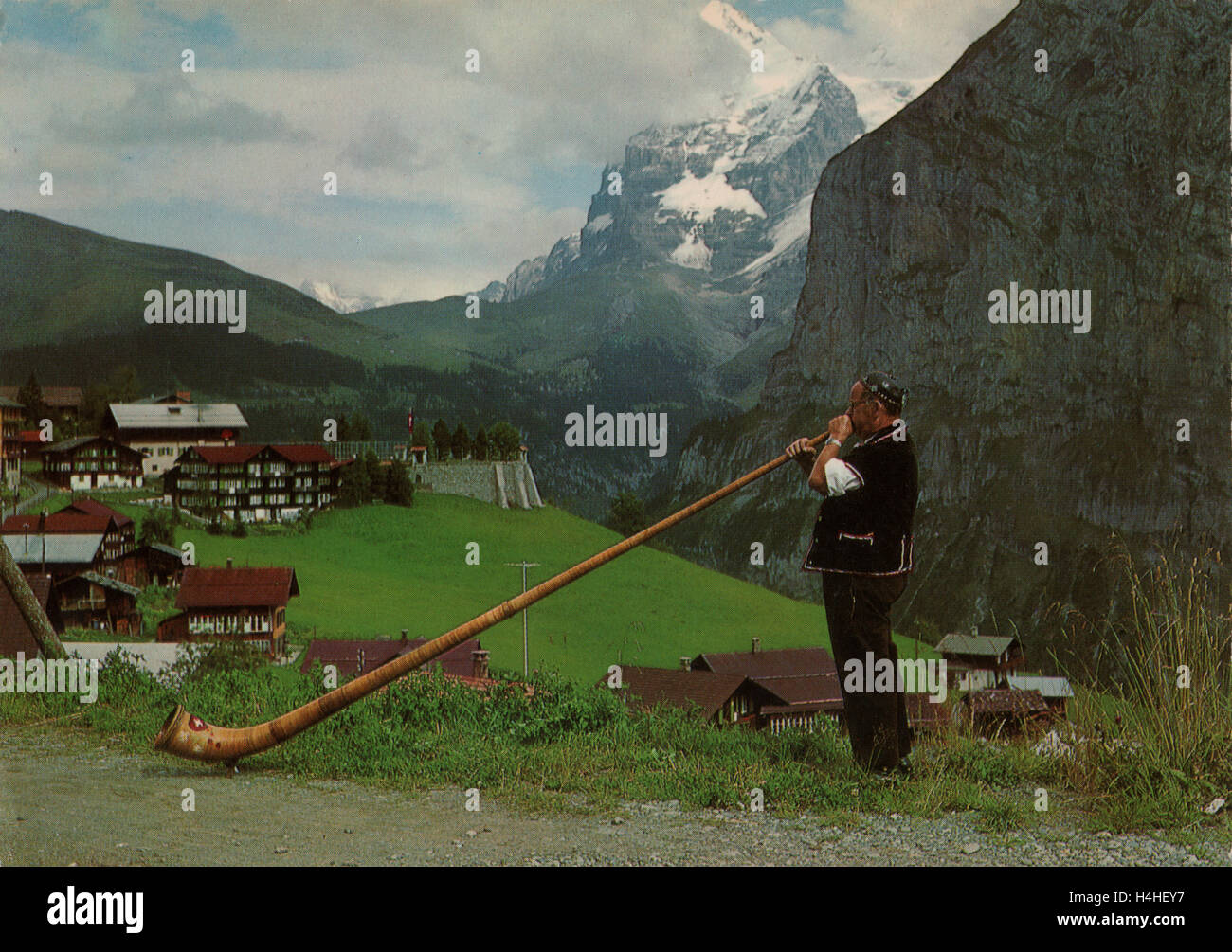 Vintage postcard of a man in traditional Swiss attire blowing an Alpine Horn or alphorn with a Swiss style mountain and rural backdrop circa 1960s. Stock Photo